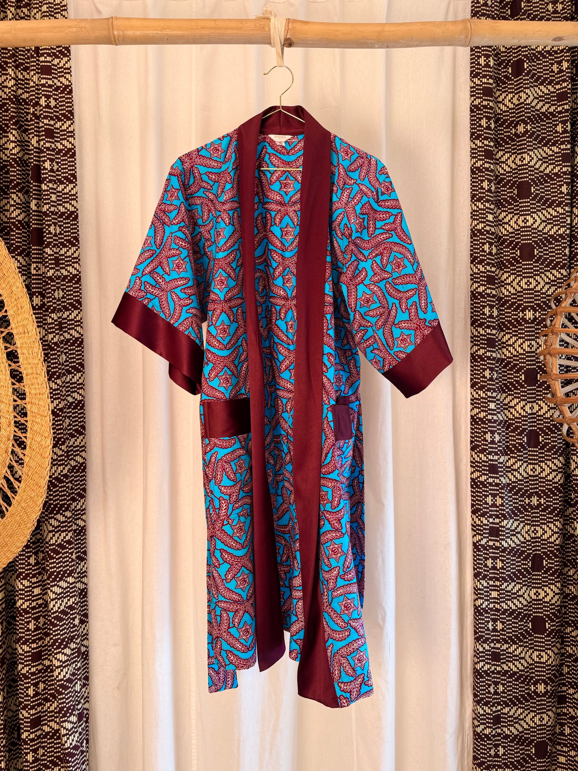 Burgundy and blue patterned robe hanging on a wooden stick against a white curtain with decorative elements.